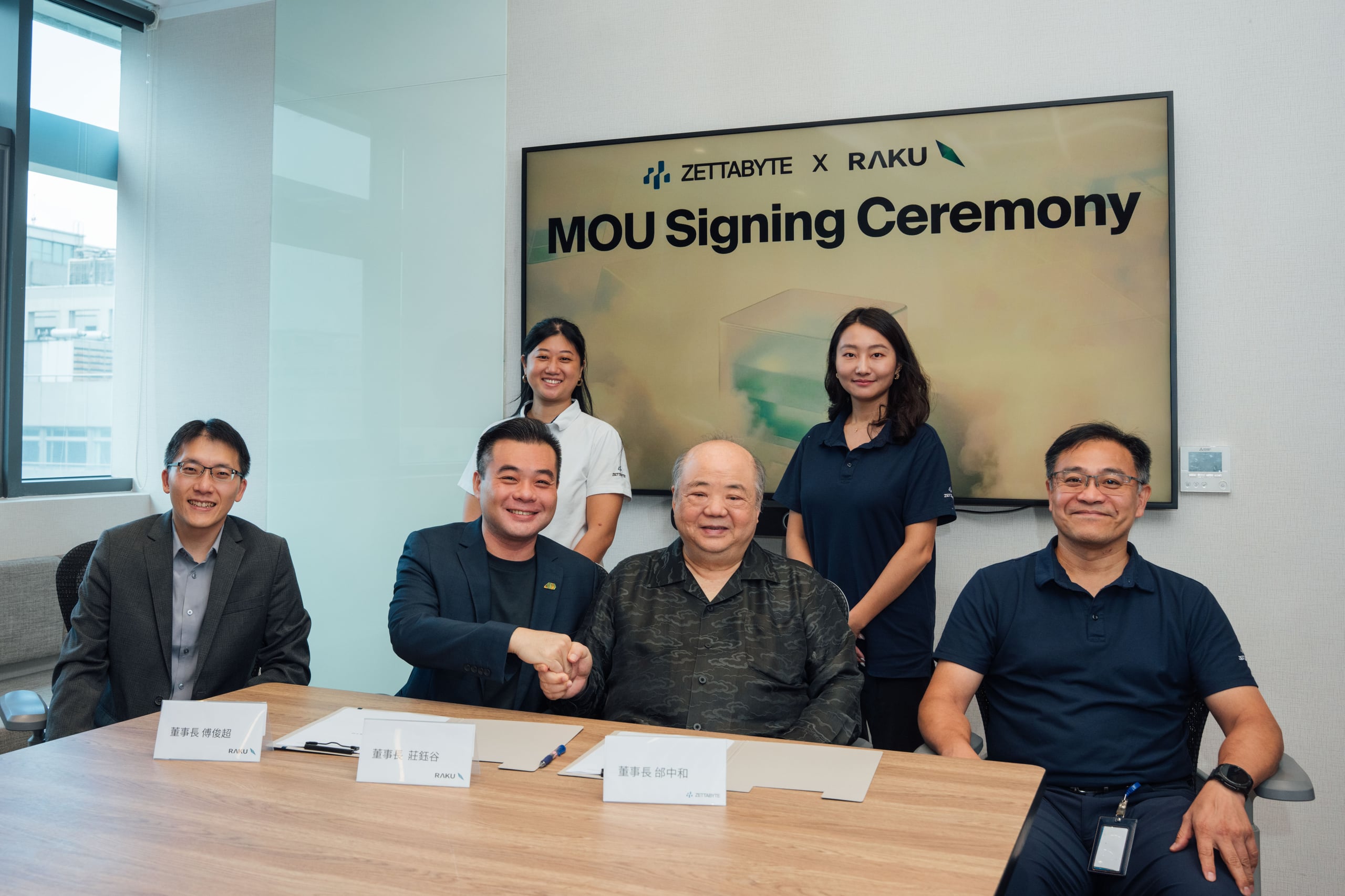 Representatives of Zettabyte and Raku Advanced Tech shaking hands across a signing table, with a screen behind them reading MOU Signing Ceremony under both company logo